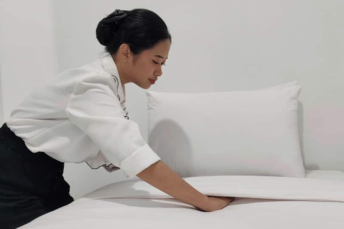 Una persona acomodando un edredón blanco sobre una cama con una almohada blanca.