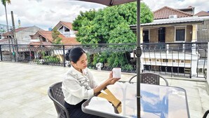 Une personne est assise à une table en plein air, d’où elle a une vue dégagée sur les bâtiments et la végétation environnants.
