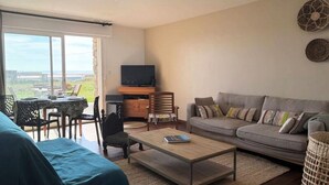 Un salon avec un canapé, une table basse et un téléviseur, un coin repas avec des chaises et une table, et un balcon avec vue.