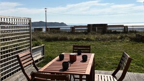 Une terrasse en bois avec une table et des chaises, donnant sur une pelouse et offrant une vue sur la mer et les collines lointaines.