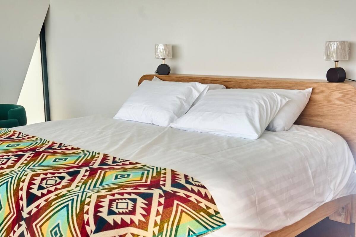 Una cama con ropa blanca y una manta colorida, dos lámparas de noche y una silla verde.