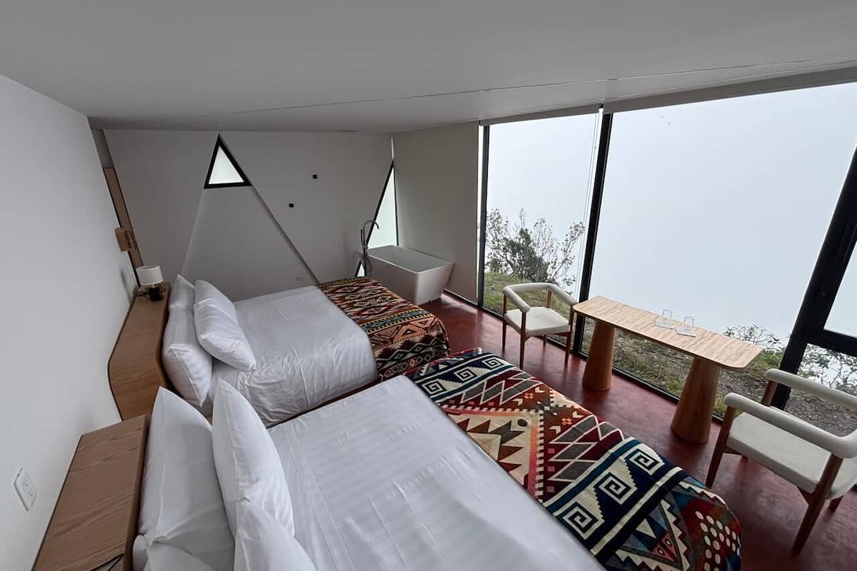 Habitación moderna con dos camas, una mesita y una silla. Un ventanal grande ofrece vistas al exterior.