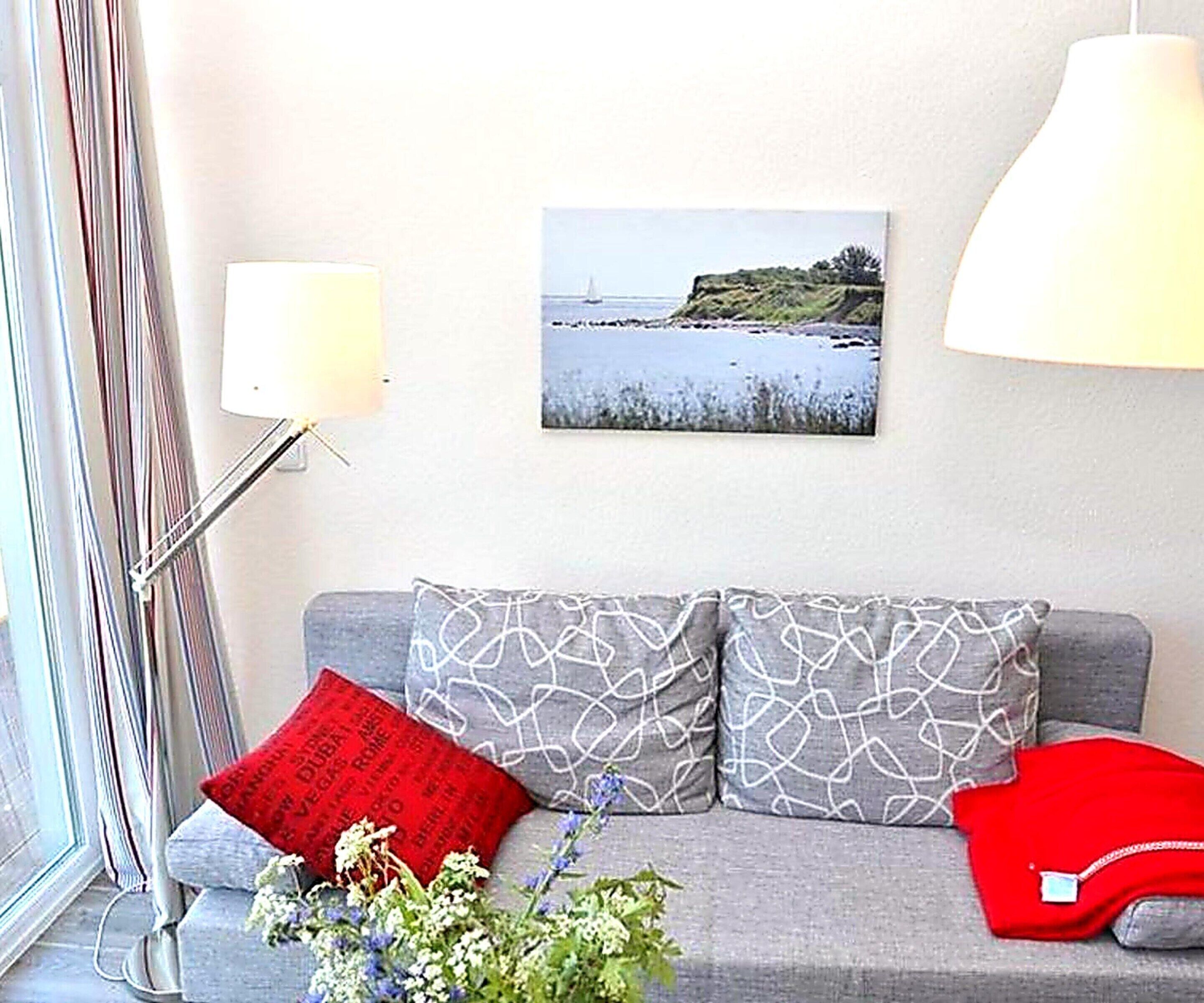 Ett vardagsrum med en grå soffa, en röd kudde, en vit lampa och en inramad bild som föreställer en strandvy.