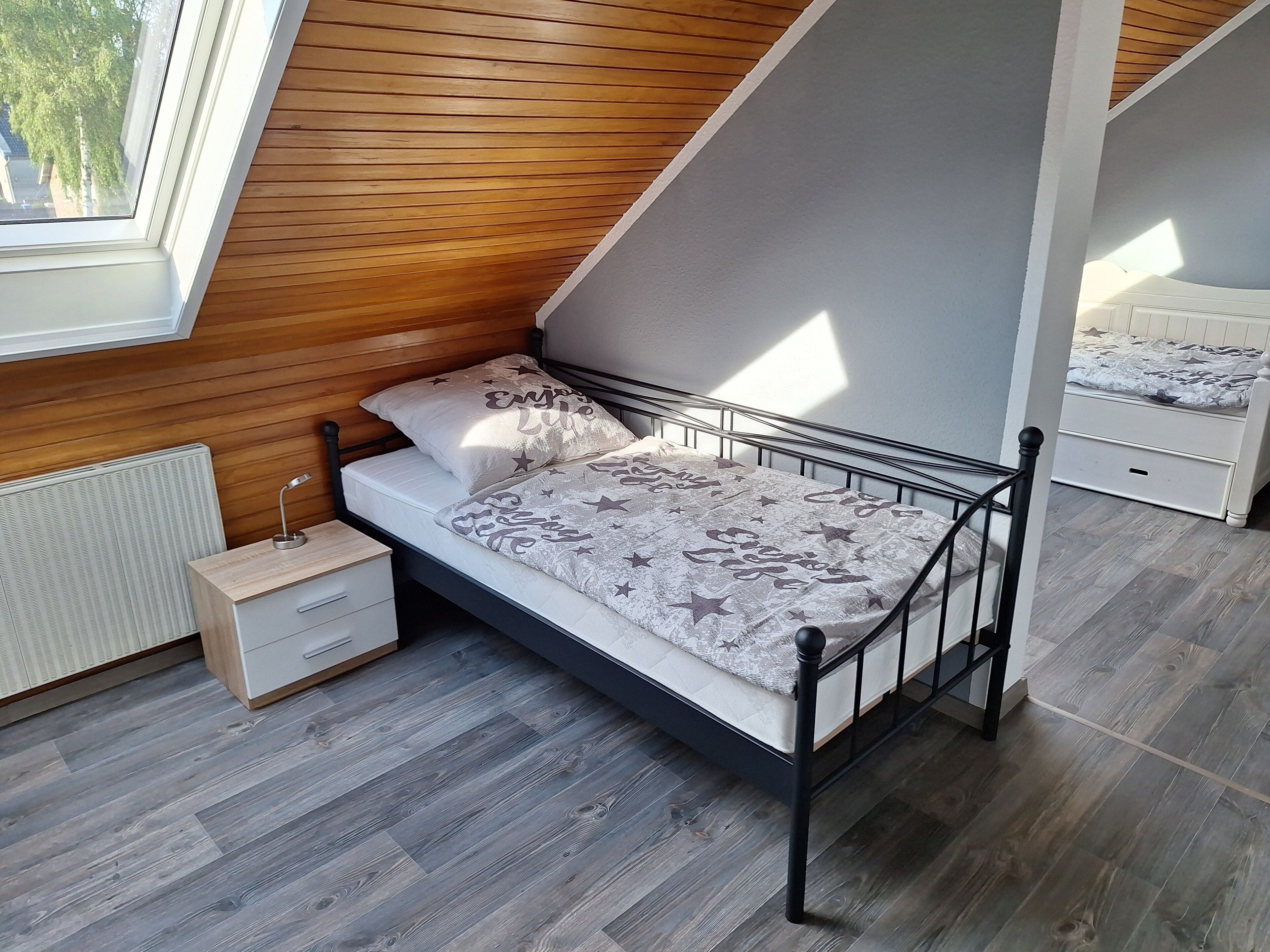 Ein Schlafzimmer mit schrägem Dach, einem Bett mit gemusterten Betttextilien, einem Nachttisch und einer Heizung.