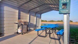 Un área de picnic techada con una parrilla, un cilindro de gas, dispensador de bolsas para excrementos de perro y mesas de picnic azules.