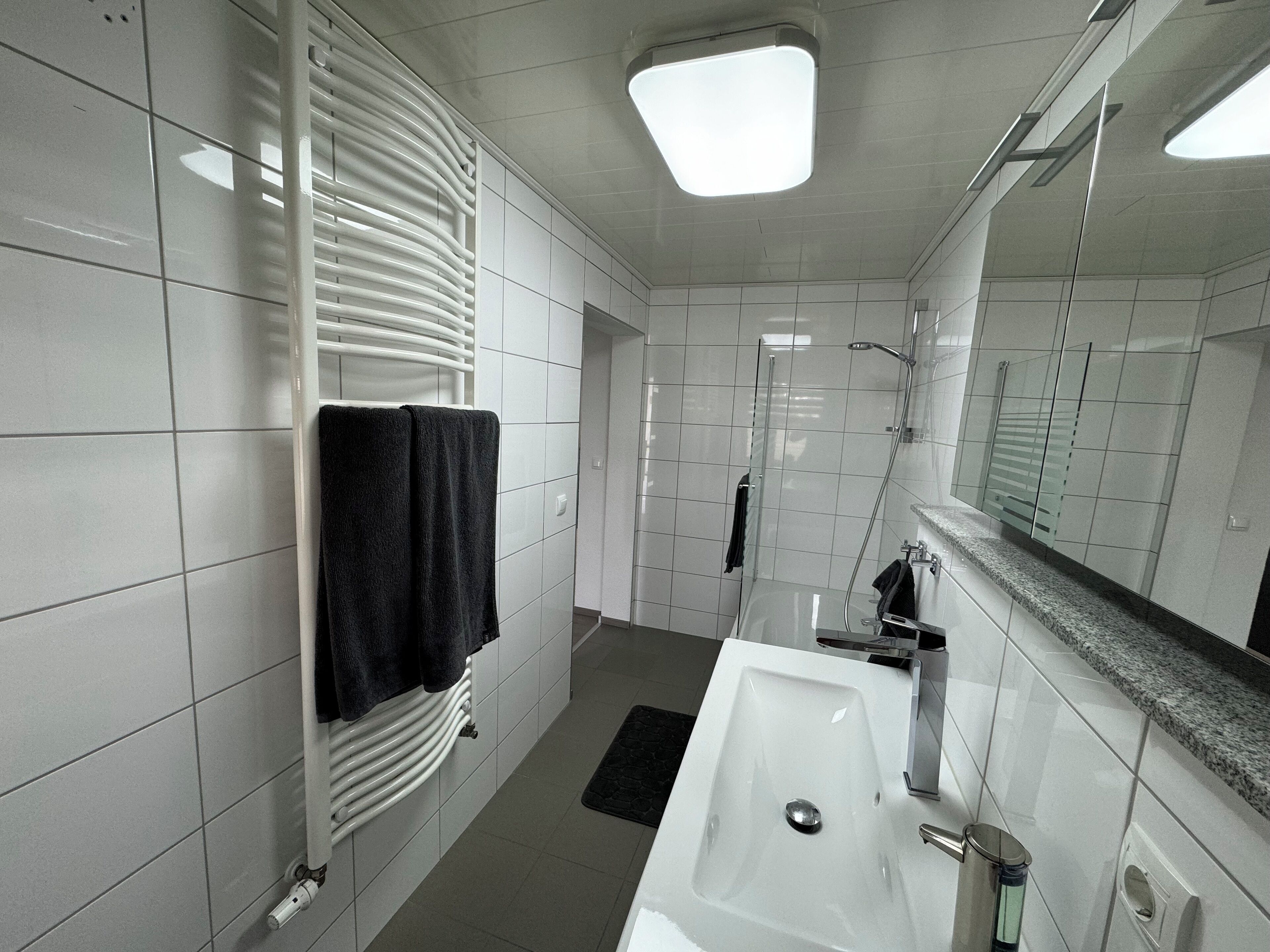 Un baño moderno con azulejos blancos, un espejo grande, un lavamanos blanco y una zona de ducha.
