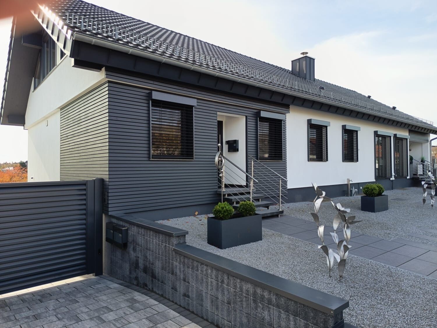 Una casa moderna con fachada gris, tejado de tejas y un pequeño jardín delantero con una escultura de metal.