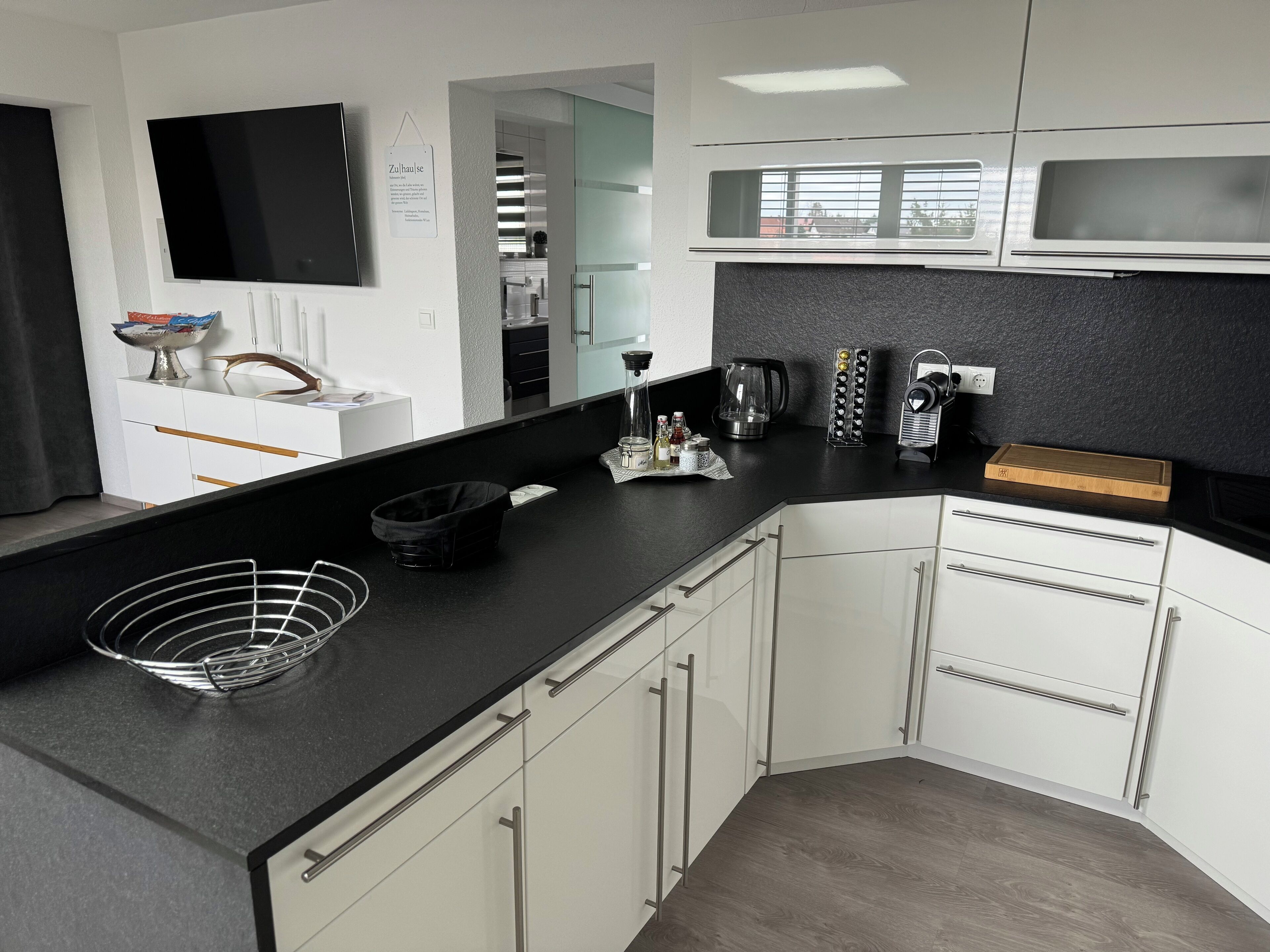 A modern kitchen with white cabinets, a black countertop, and a flat-screen TV mounted on the wall.