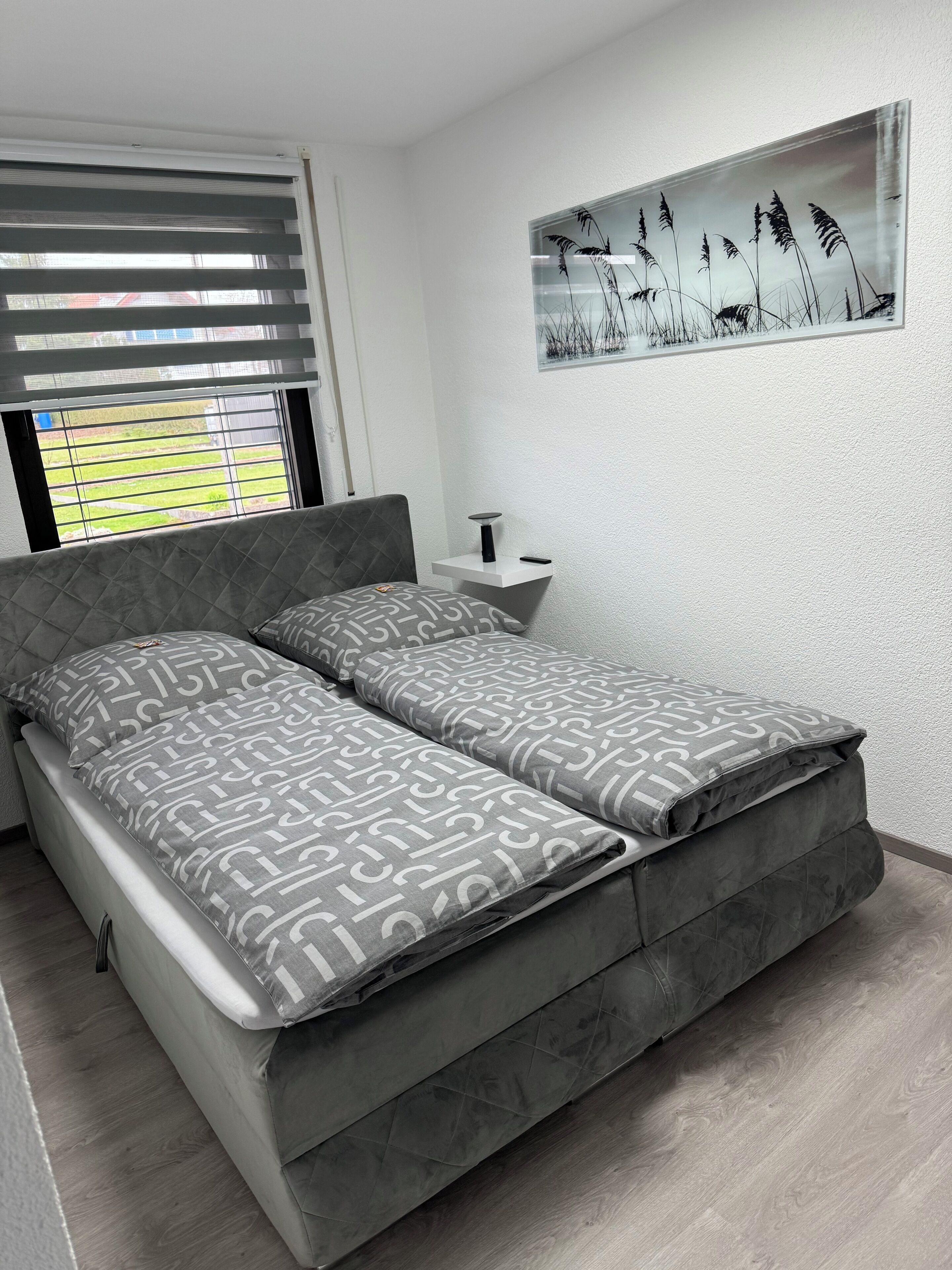 Un dormitorio moderno con una cama gris, una mesita de noche pequeña y una obra de arte en la pared que representa pastos altos.