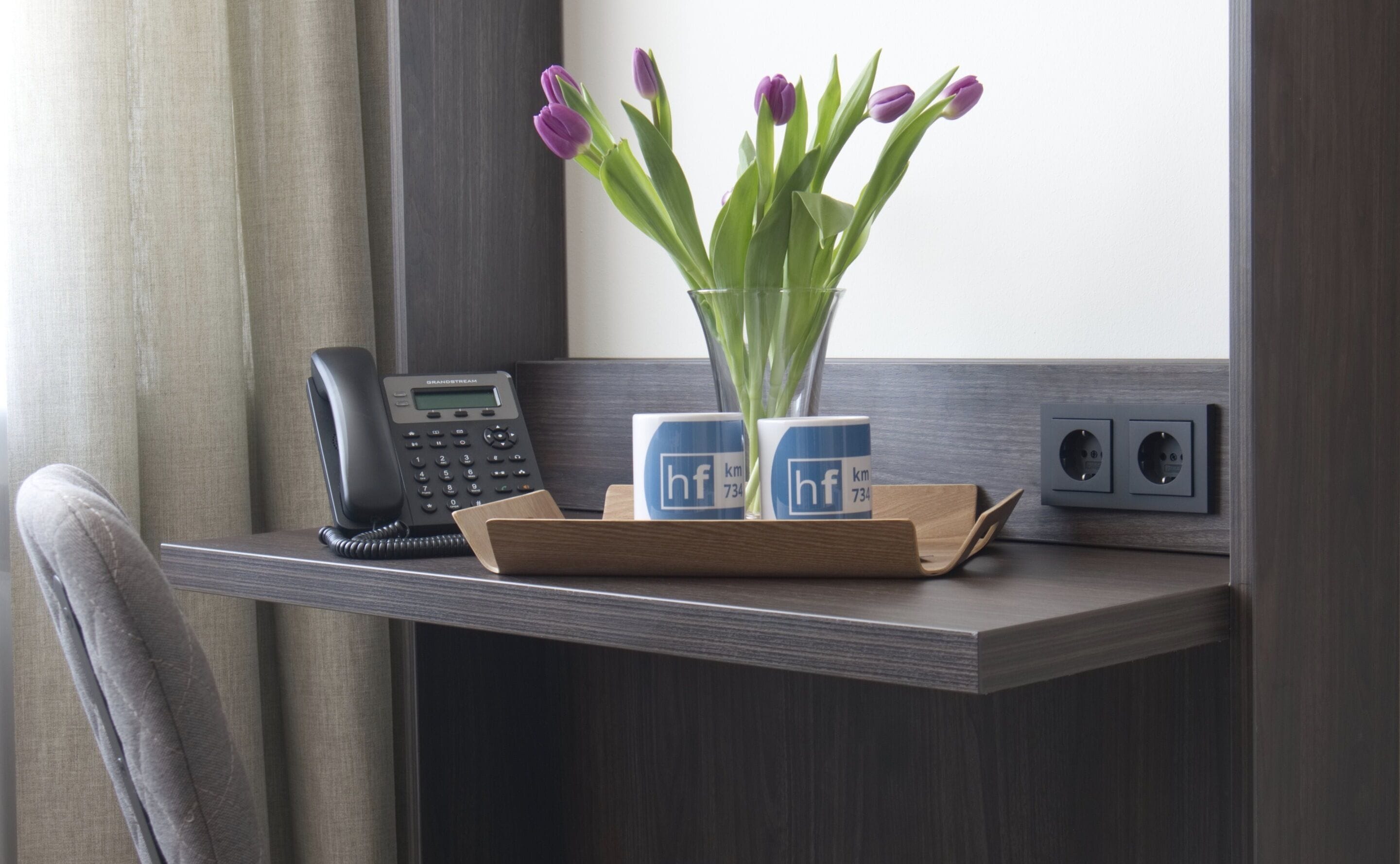 A desk with a telephone, two branded coffee cups, a vase with purple tulips, and a power outlet.