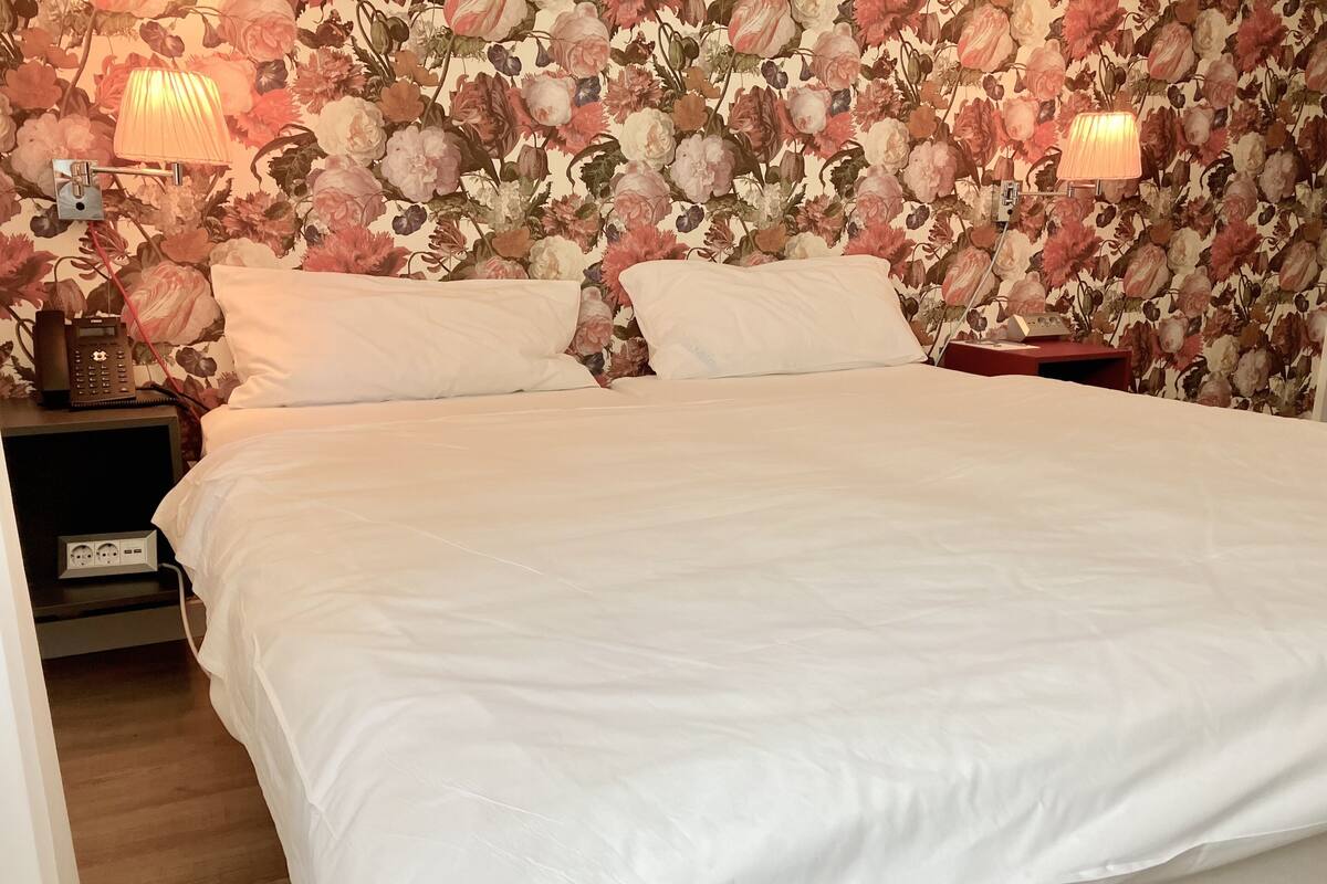 A neatly made bed with white linens, two bedside lamps, and a floral wallpaper.