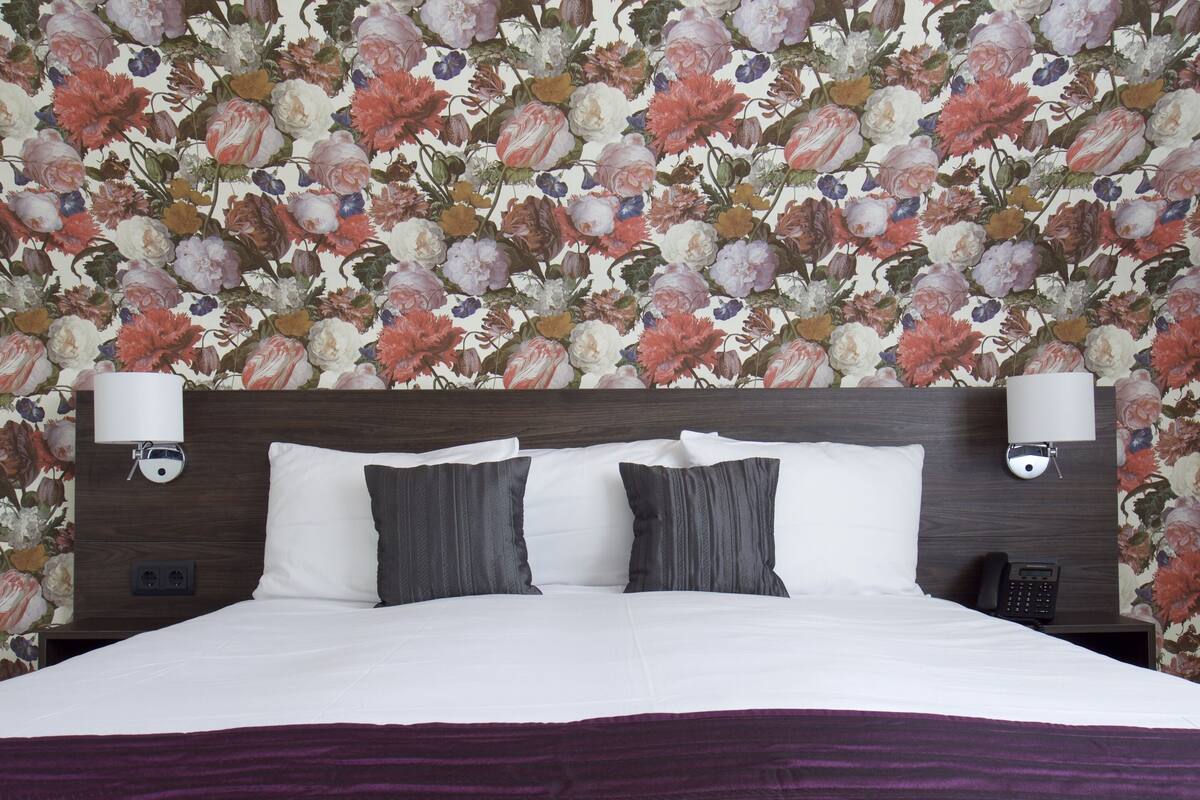 A hotel room with a floral wallpaper, a wooden headboard, two bedside lamps, a telephone, and a purple bedspread.