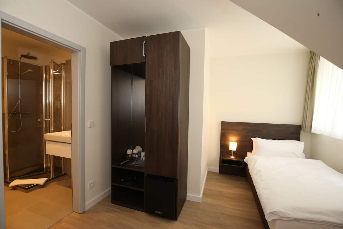A hotel room with a walk-in closet, a shower, a bedside table with a lamp, and a bed.