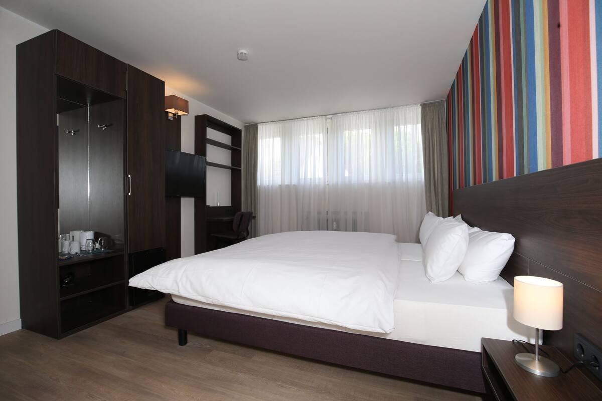 A hotel room with a large bed, a wardrobe, a desk with a chair, and a TV.