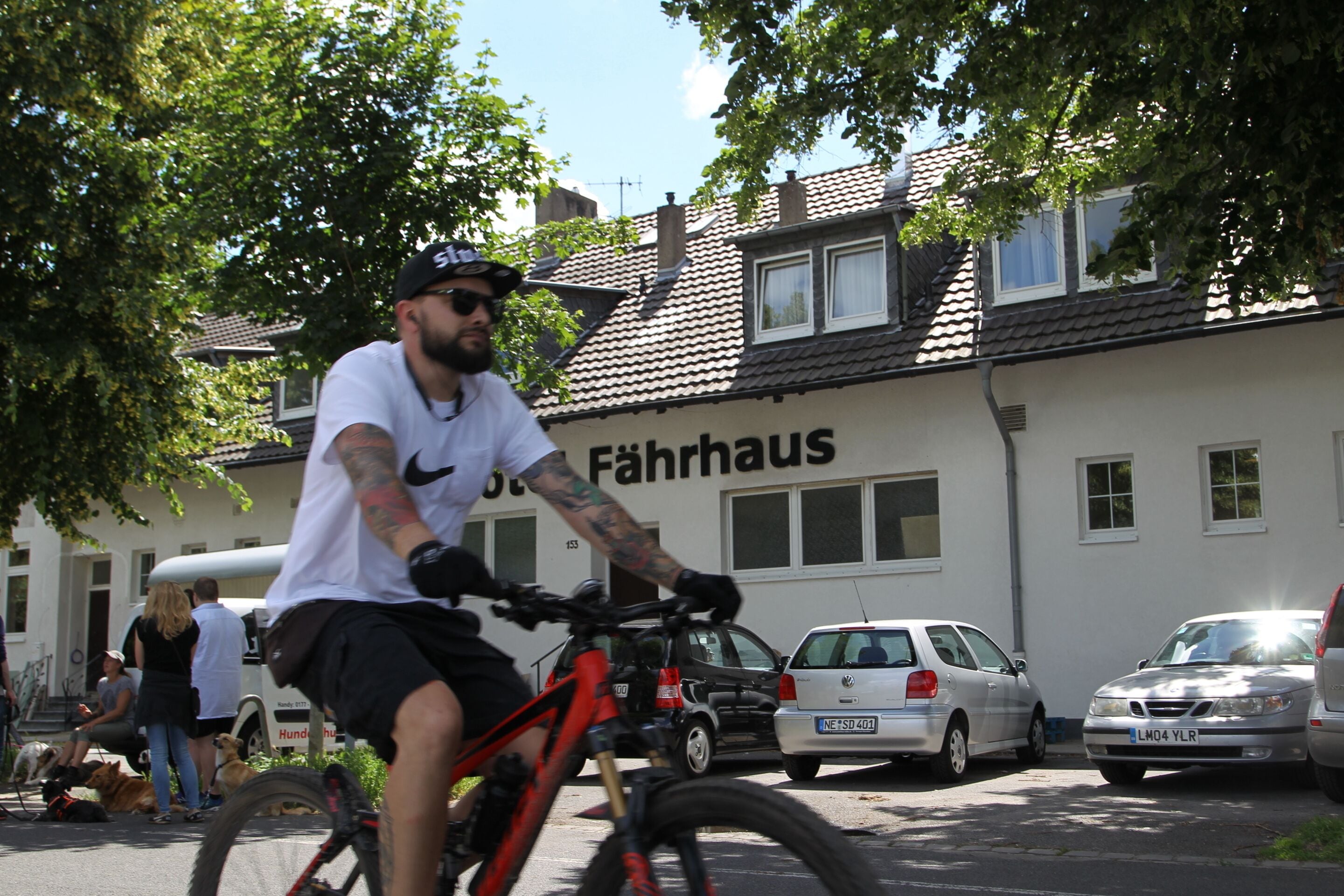 A person riding a bicycle on a street with parked cars and a building labeled "Fahrhaus" in the background.