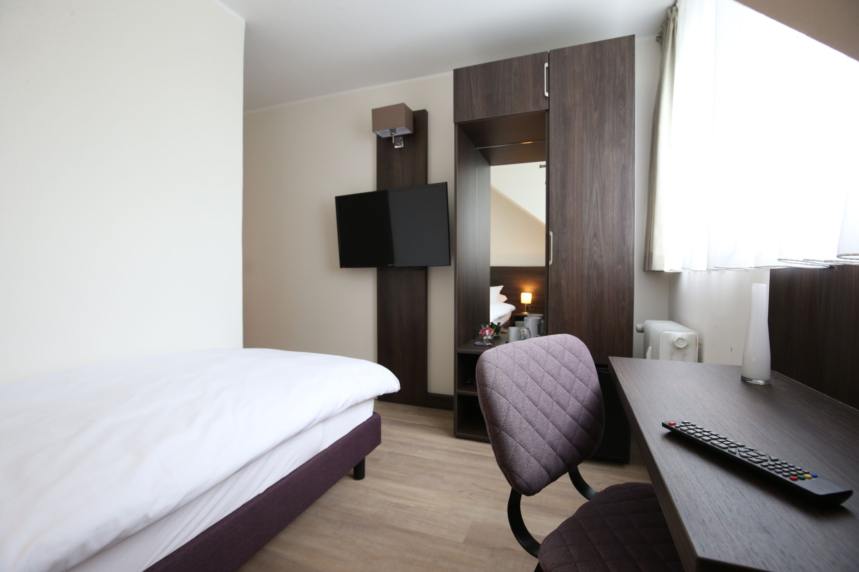 A hotel room with a bed, a desk, a chair, a television mounted on the wall, and a window with curtains.