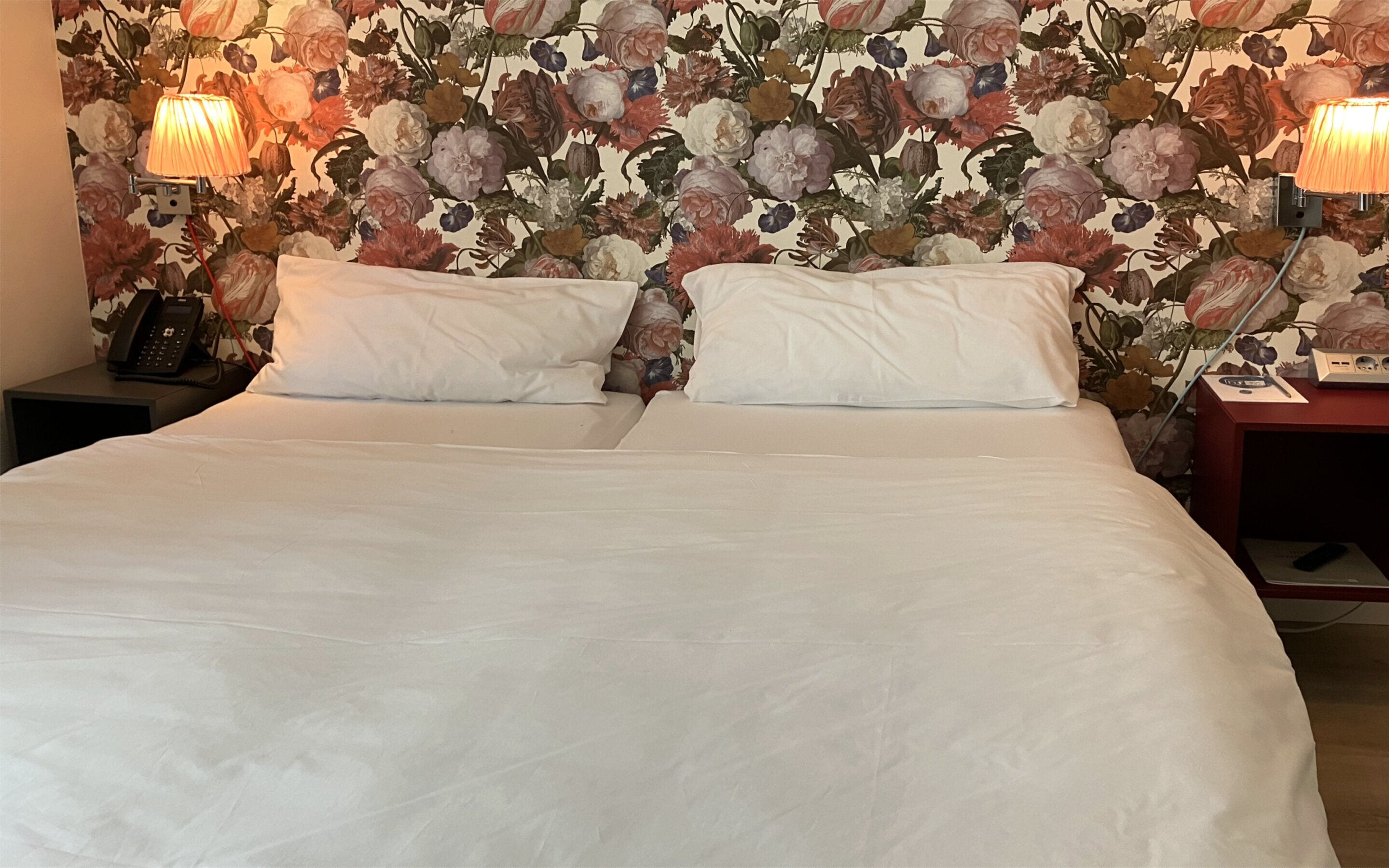 A neatly made bed with white linens, two bedside lamps, and a floral wallpaper.