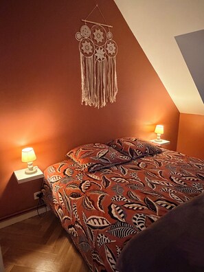 A bedroom with a sloped ceiling, a patterned bedspread, two bedside lamps, and a wall hanging.