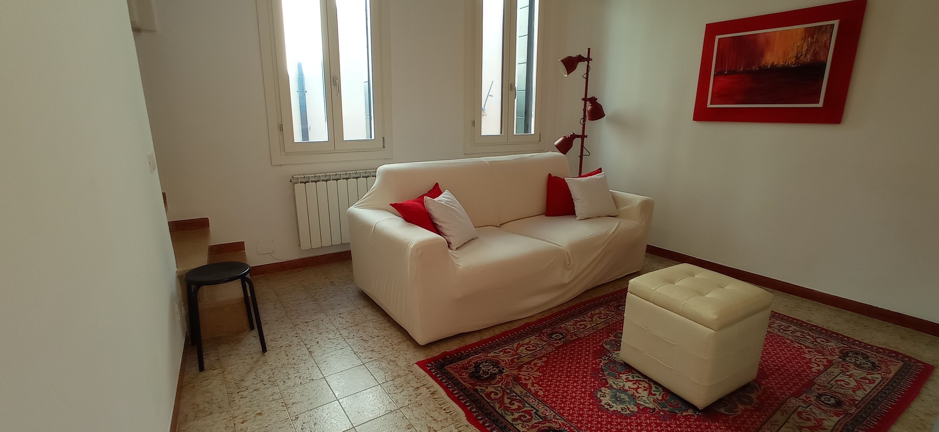 A living room with a white sofa, red cushions, a small black chair, a red and beige rug, and a floor lamp.