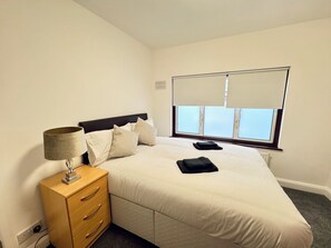 Ein Hotelzimmer mit einem Bett, einem Nachttisch, einer Lampe, einem Fenster mit Jalousien und Blick auf den Himmel.