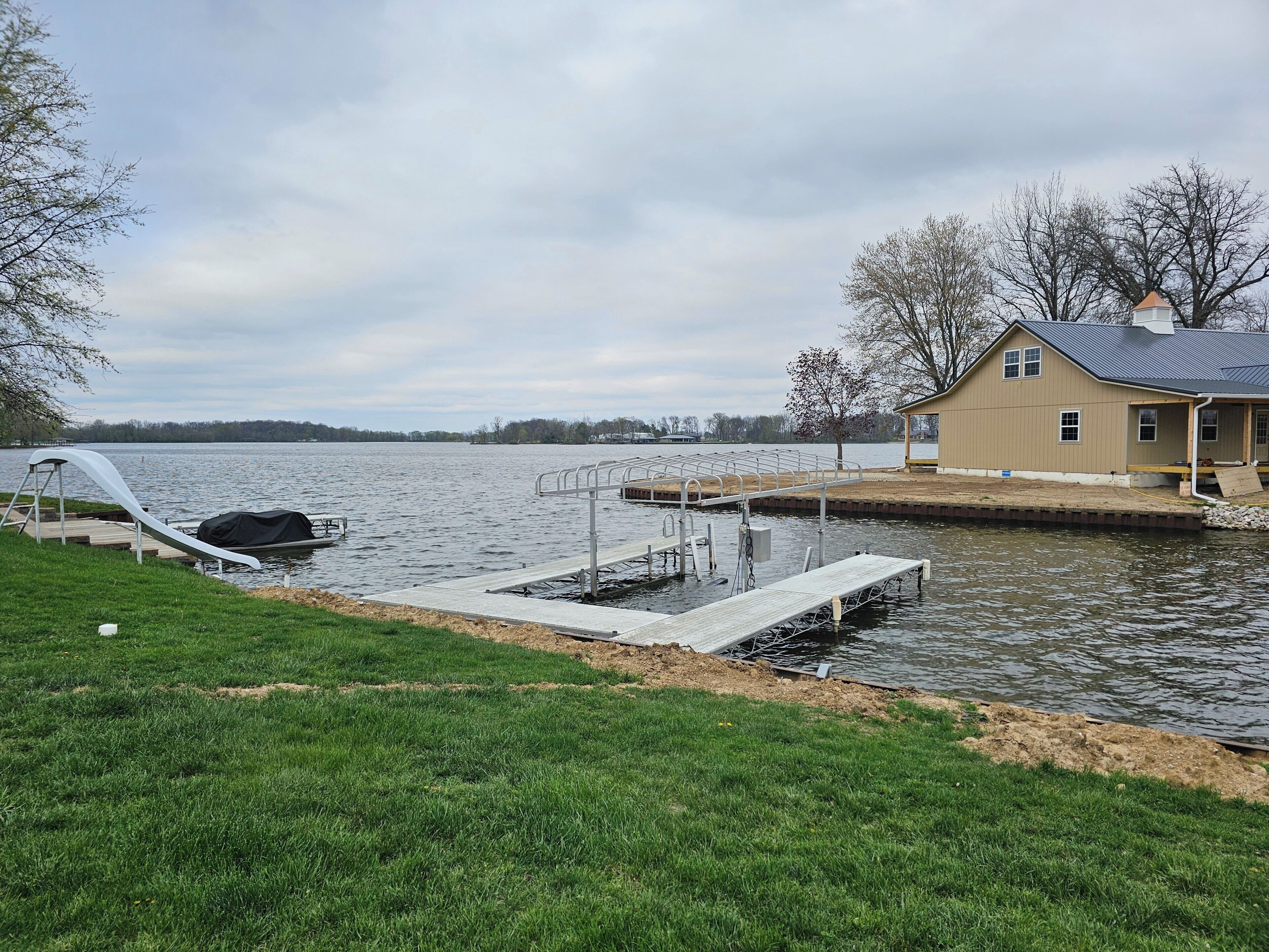 A lakeside dock with a covered boat, a house with a porch, and a grassy area.