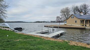 A lakeside dock with a covered boat, a house with a porch, and a grassy area.