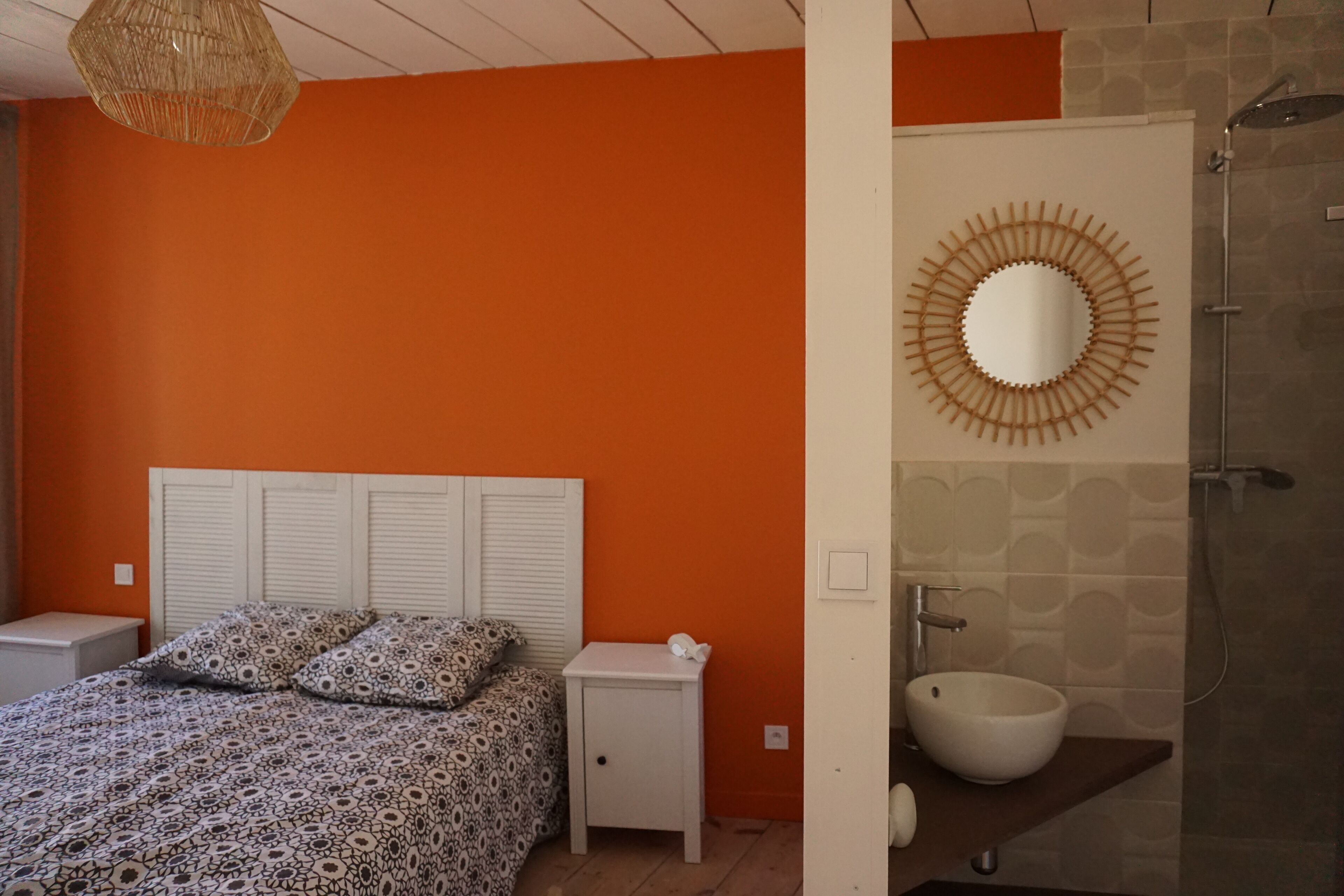 A bedroom with a bed, bedside tables, a nightstand, a hanging light fixture, and a bathroom with a sink and mirror.