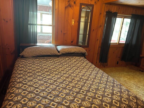 A wooden cabin bedroom with a patterned bedspread, two pillows, a wooden headboard, and a window with curtains.