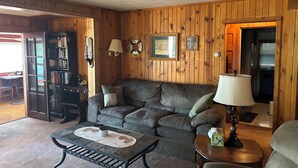 A cozy living room with a wooden paneled wall, a grey sofa, a coffee table, and a bookshelf. - Beautiful one acre 3 bedroom Private boat and snowmobile launch (Saint Germain)