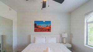 A bedroom with a large bed, white bedding, a nightstand with a lamp, a painting of a desert landscape, and a window with blinds. - San Portella 2019 (Tempe)