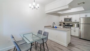A modern kitchen with white cabinetry, a stainless steel refrigerator, a microwave, and a gas stove. There is a glass dining table with four chairs. - San Portella 2019 (Tempe)
