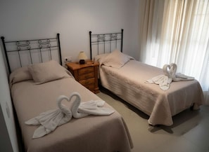 Dos camas individuales con cabeceras y pies de cama a juego, una mesita de noche de madera con lámpara, y una ventana con cortinas de gasa.