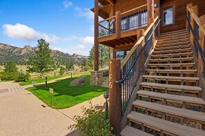 A wooden cabin with a staircase leading to a balcony, surrounded by a well-maintained lawn and mountains in the background. - Stunning Lumpy Ridge Views • Near RMNP & Downtown (Estes Park)