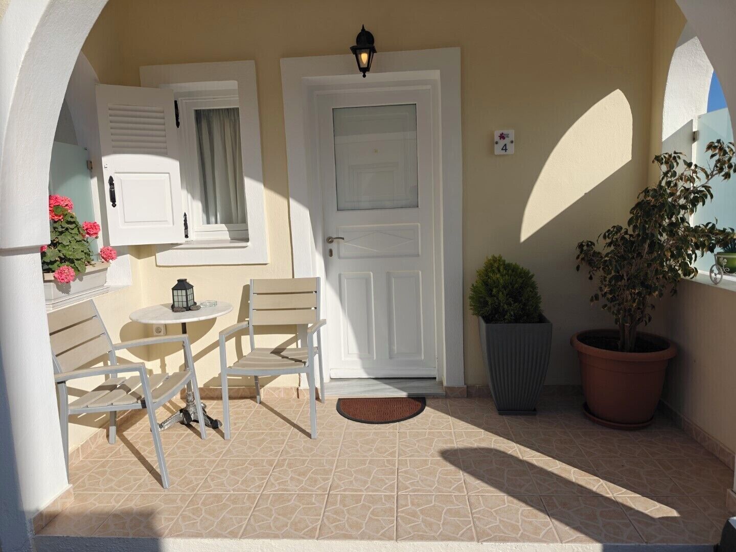 A small outdoor patio with a table and chairs, a door, a window with shutters, and potted plants.