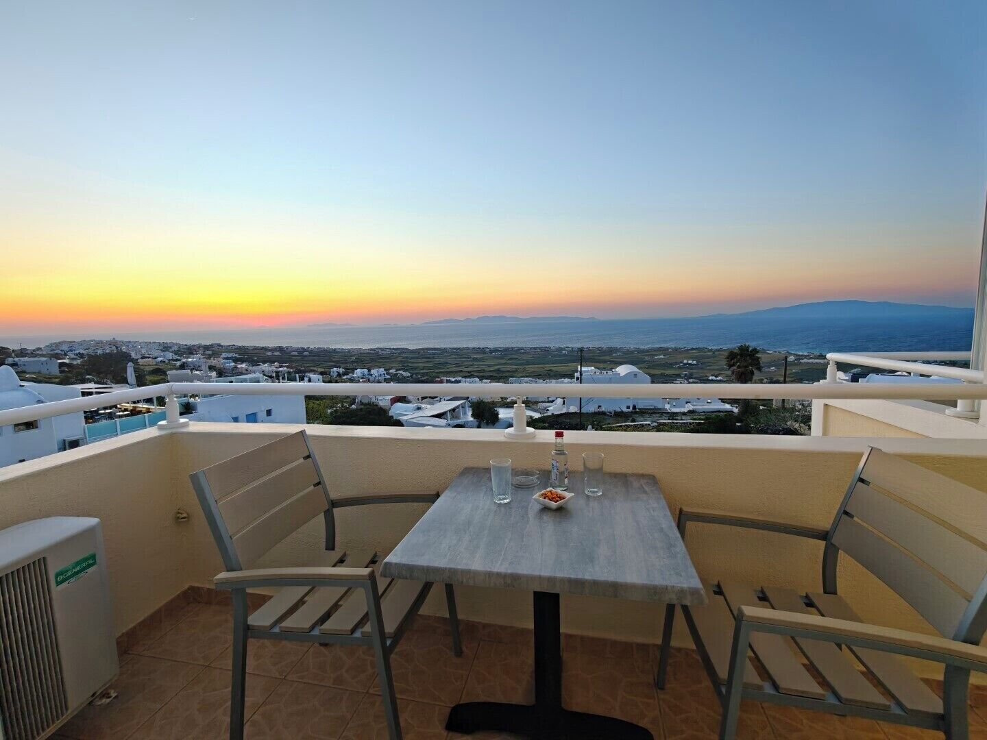 A balcony with a table and chairs, overlooking a coastal landscape with buildings and a sunset.