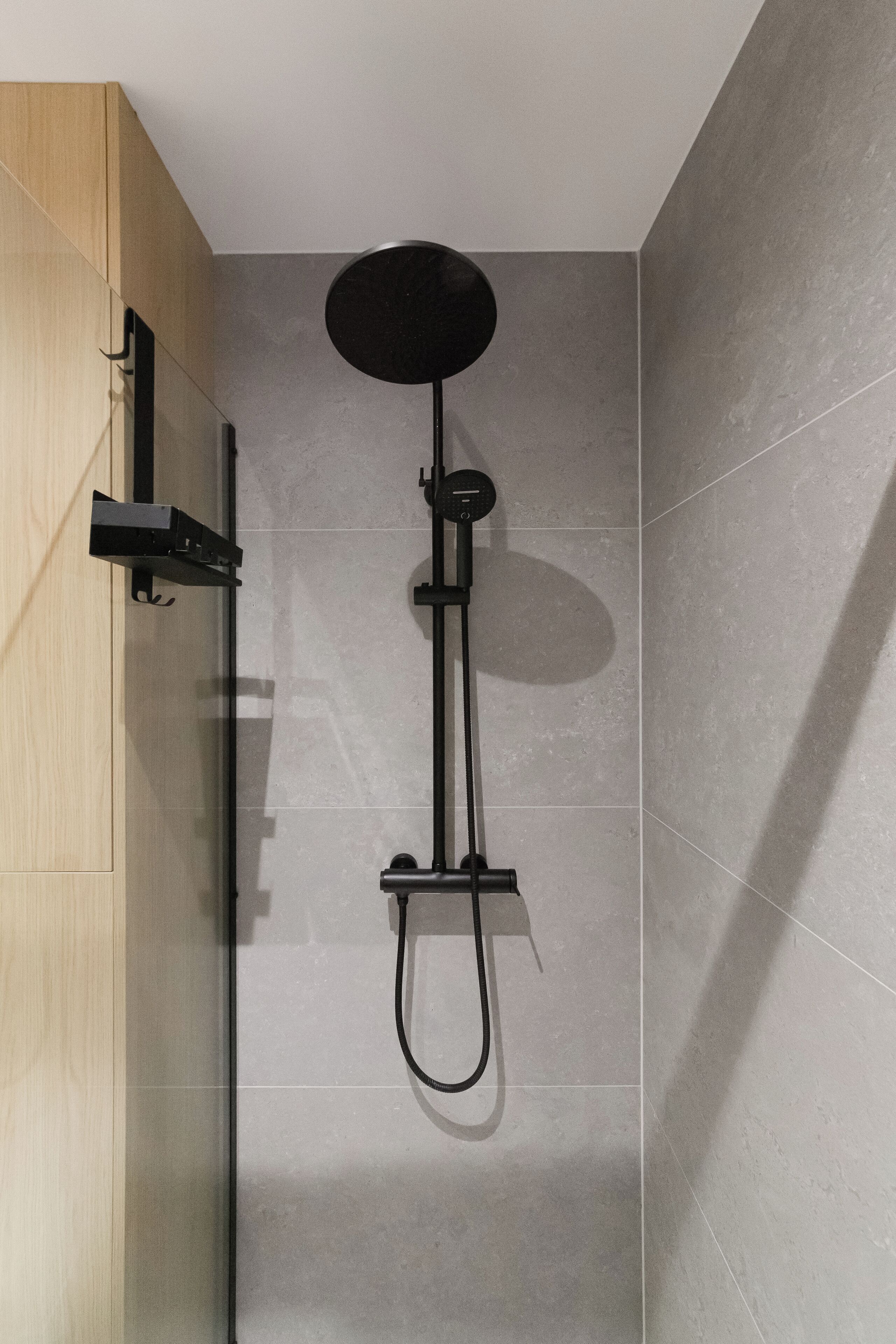A modern shower with a black head and a handheld showerhead, mounted on a wall with grey tiles.