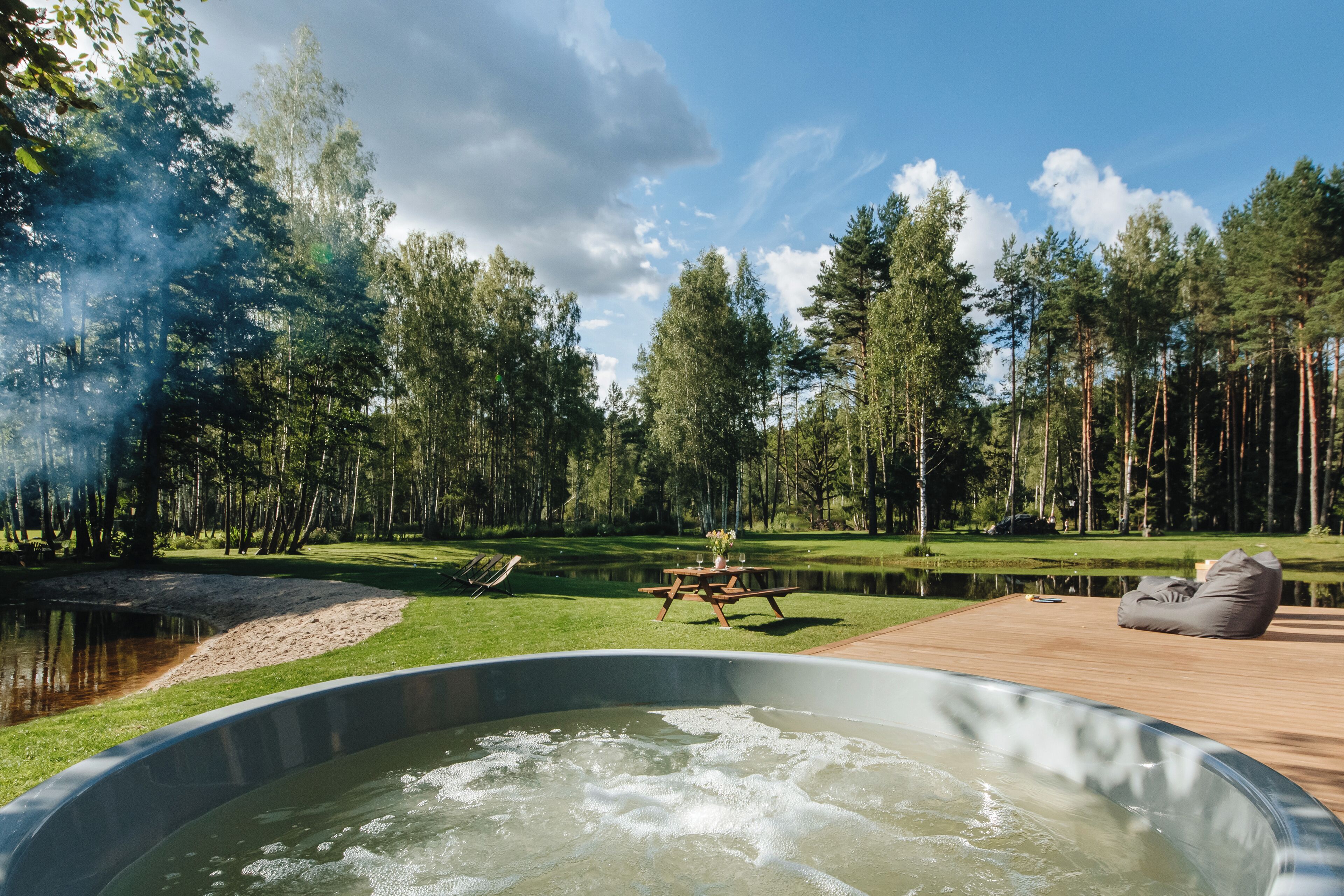 A hot tub on a wooden deck with a picnic table and bean bag chairs, surrounded by a lake and a forest.
