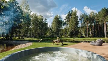 A hot tub on a wooden deck with a picnic table and bean bag chairs, surrounded by a lake and a forest.