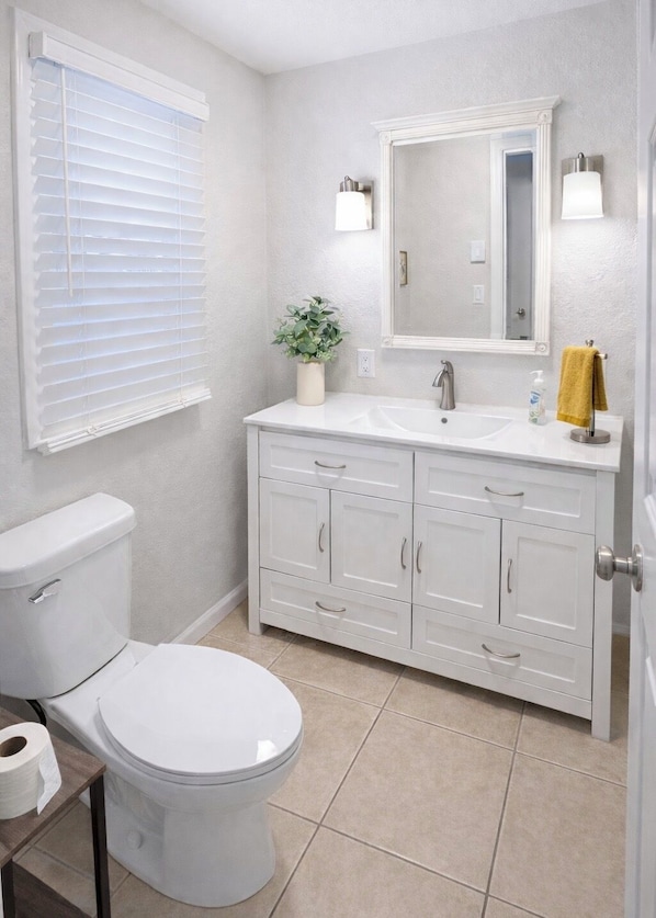 A bathroom with a toilet, sink, cabinet, mirror, and window with blinds.