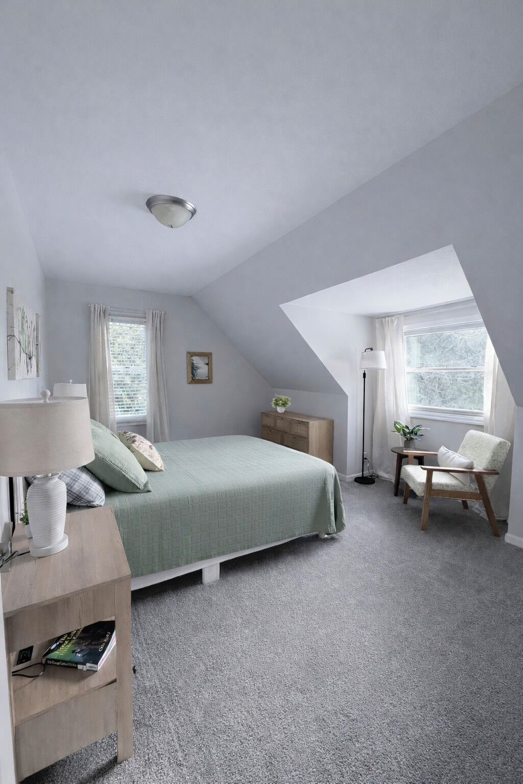 Une chambre avec un plafond en pente, un lit recouvert d’une couverture verte, une table de chevet avec une lampe, une chaise, une petite table avec une plante et une fenêtre avec des rideaux.