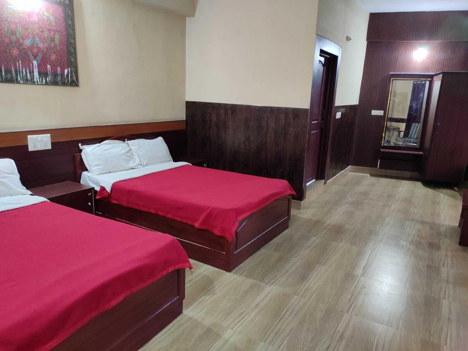 A hotel room with two beds, a wooden headboard, a red bedspread, a wooden floor, and a painting on the wall.