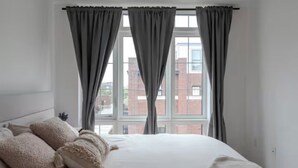 A bedroom with a large bed, grey curtains, and a view of a brick building through the window. - Quiet charming townhome condo (Whitchurch-Stouffville)