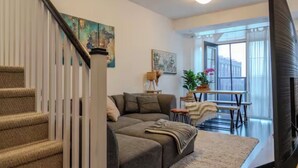 A modern living room with a staircase, a sofa, a dining table, and a view of the city through large windows. - Quiet charming townhome condo (Whitchurch-Stouffville)