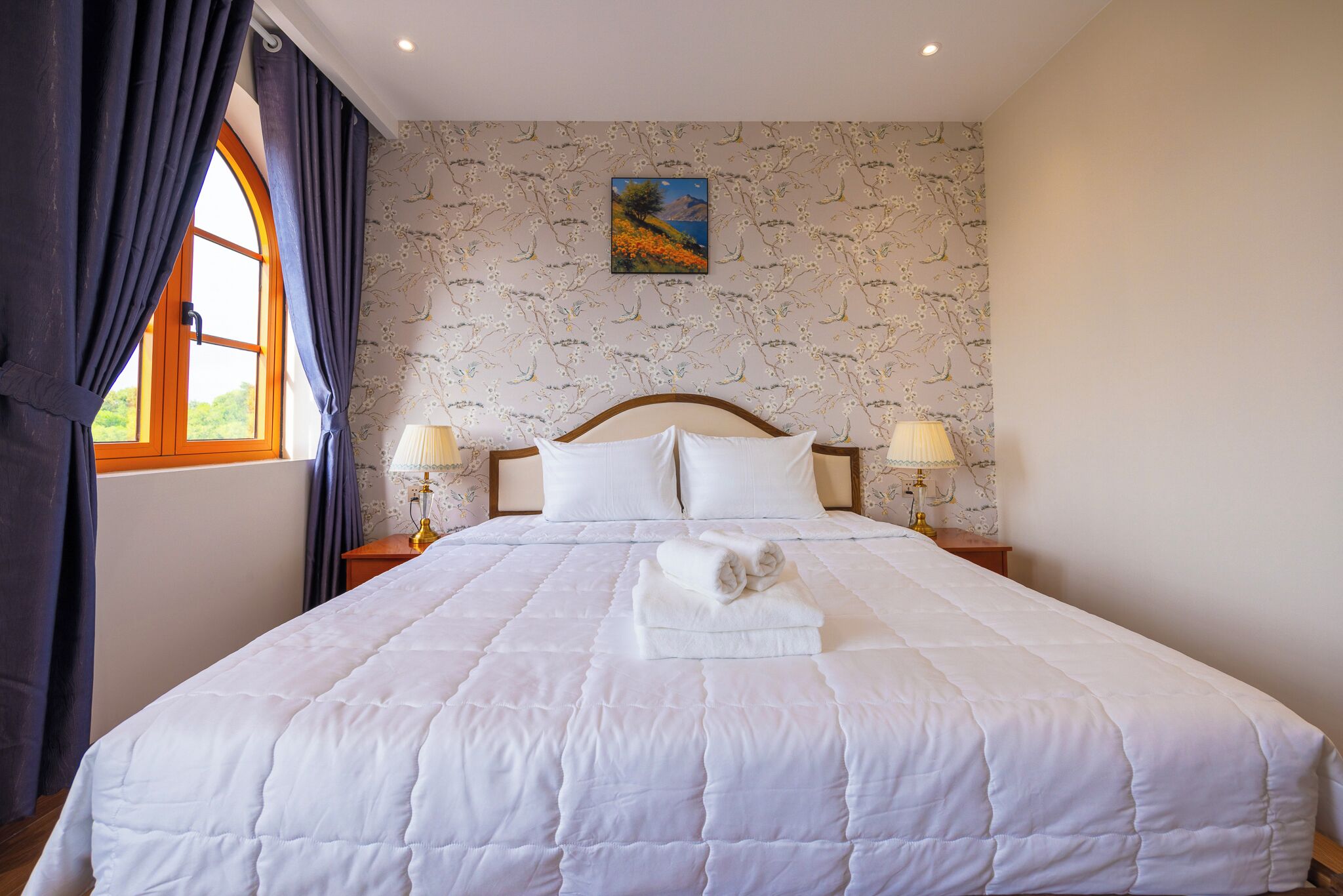 A neatly made bed with white linens and a folded towel, a nightstand with a lamp, and a window with curtains.