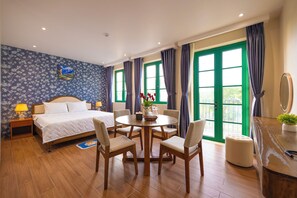 A hotel room with a bed, a round dining table with chairs, a small stool, a nightstand, a lamp, a vase with flowers, and a window with curtains. - CIEL ROSE HOTEL (Phu Quoc)