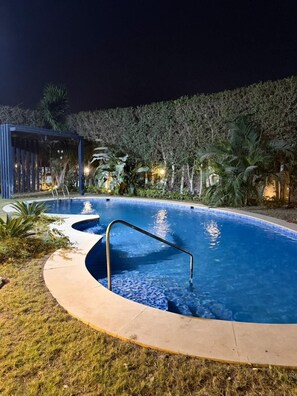 A swimming pool with a curved edge, surrounded by grass and palm trees, and a wooden structure in the background.