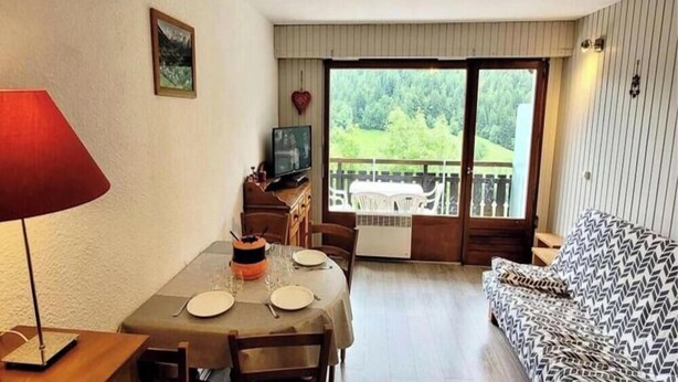 Un coin repas avec une table dressée pour deux personnes, un téléviseur posé sur un meuble en bois et un balcon offrant une vue sur la verdure.