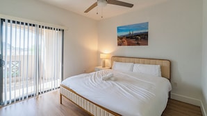 Um quarto com uma cama, um ventilador de teto, uma mesa de cabeceira com abajur, uma janela com persianas e um quadro na parede.