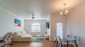 Uma sala de estar espaçosa com uma mesa de jantar de vidro, um sofá, um ventilador de teto e uma TV fixada na parede.