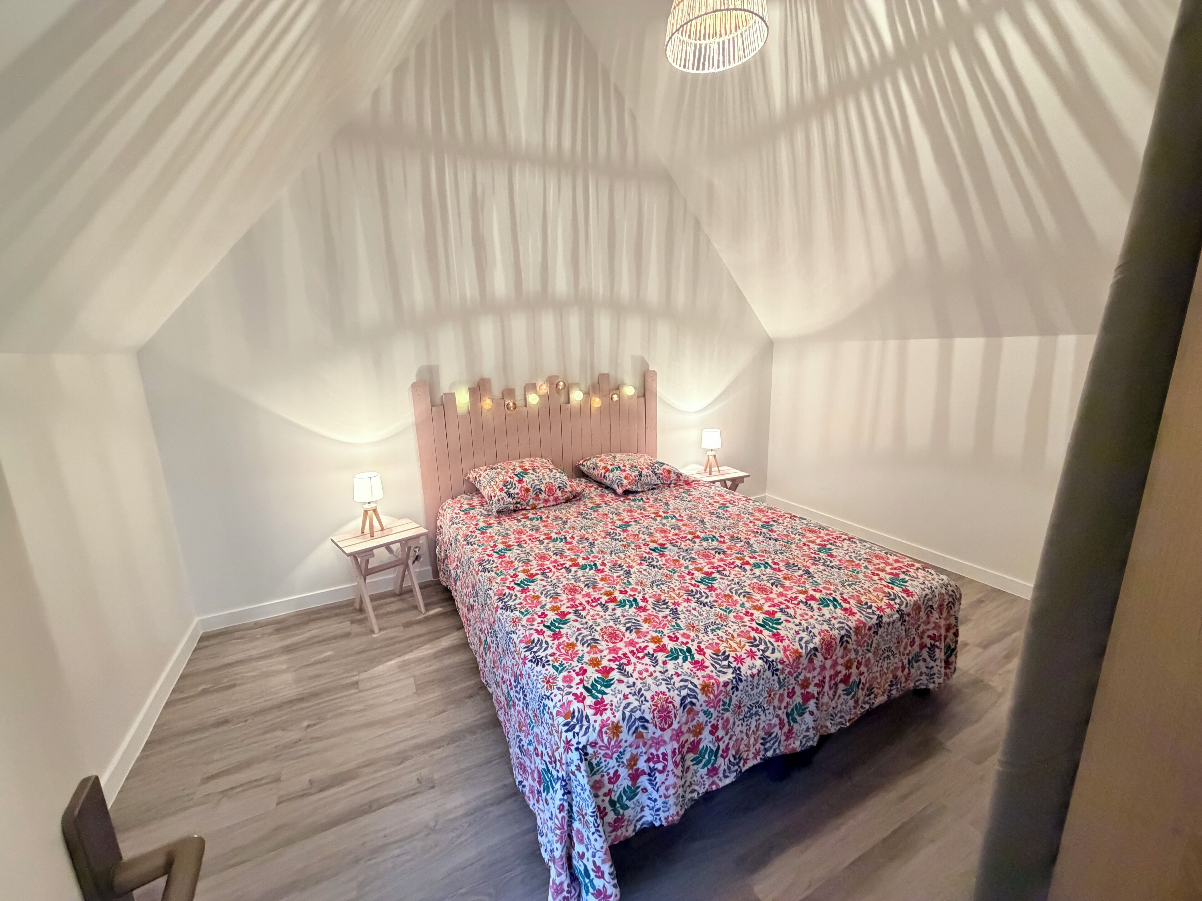 A bedroom with a sloped ceiling, a wooden headboard, a floral bedspread, a bedside table with a lamp, and a wooden floor.